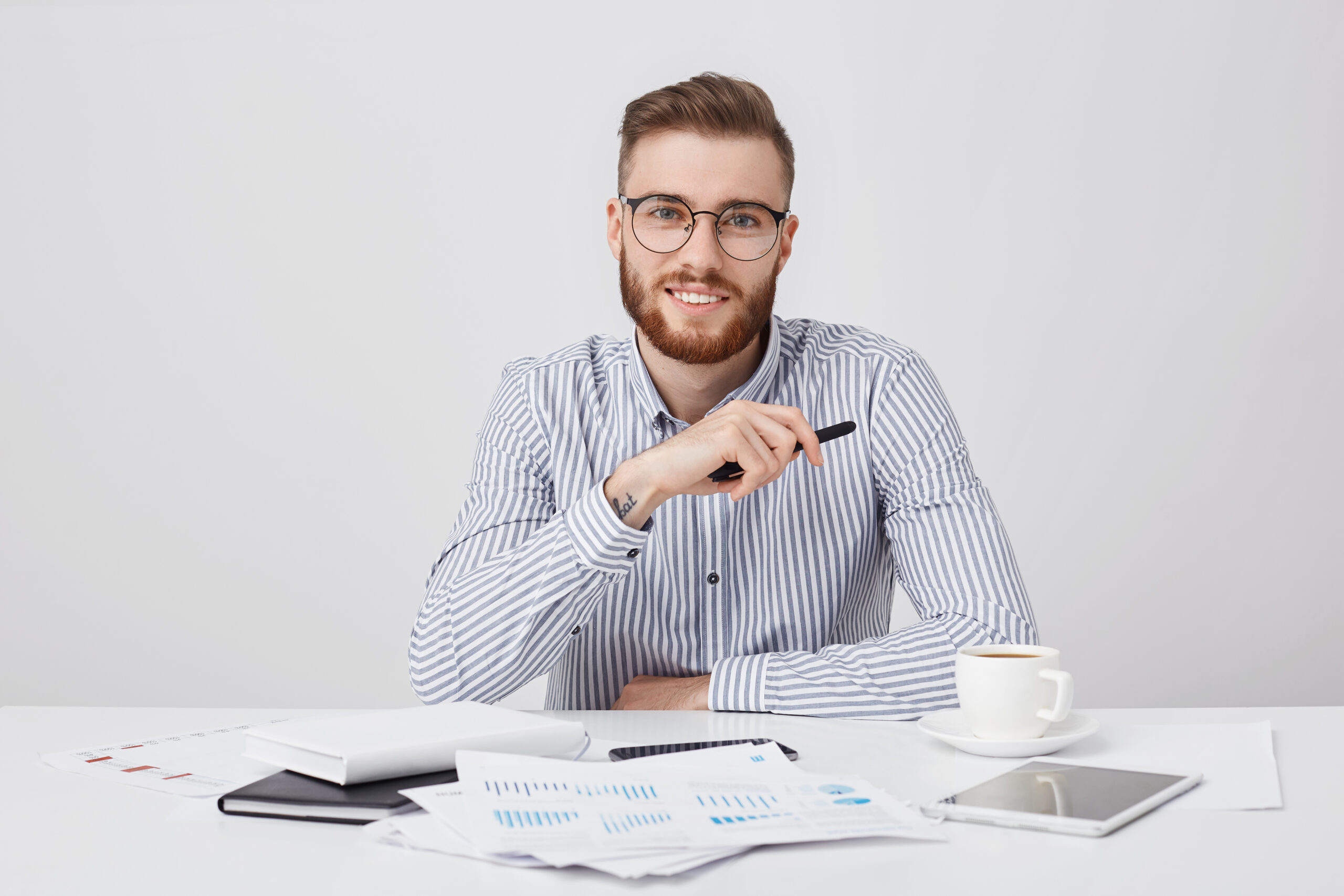 Professional male worker with thick beard and trendy hairstyle, wears round glasses and formal shirt, sits at work plae, drinks coffee, works with documents. Hipster young guy being busy working Usługi graficzne, social media i strony internetowe